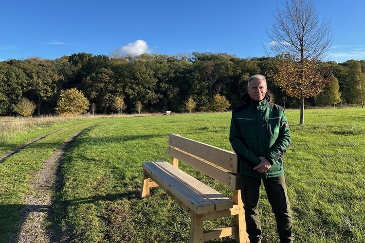 A man in a green coat stood next to a bench with trees along the horizon. 